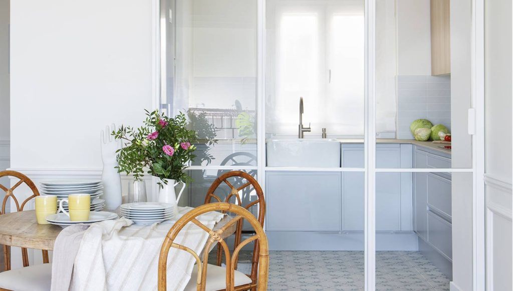 Sala de estar luminosa y decorada. De fondo, una cristalera con estructura metálica blanca transparente que deja ver una pequeña cocina recién diseñada