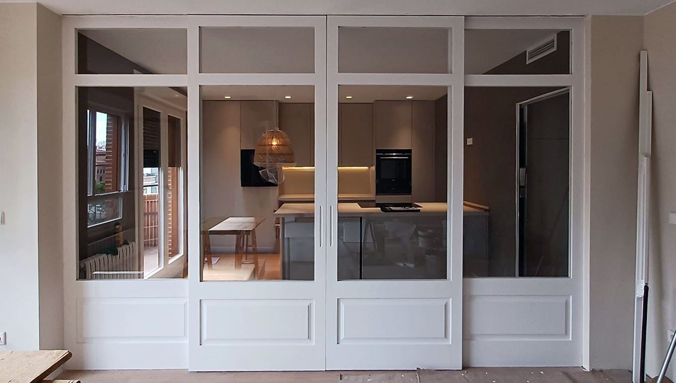 Cerramiento interior de madera blanca con cristalera. De fondo, una cocina de concepto abierto