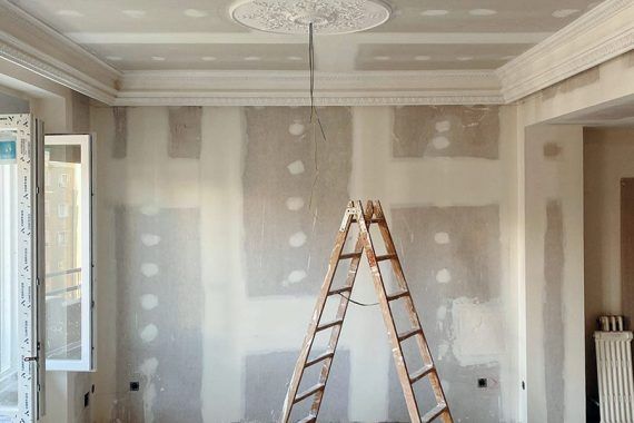 Habitación en proceso de reforma integral, con una escalera y paredes de pladur al fondo. Arriba, se puede observar un techo con molduras de escayola.